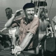 Pete Seeger aboard the sloop Clearwater during its maiden voyage, 1970. Seeger's shirt sleeves are rolled up and he's reaching hig hand up towards his dark cap